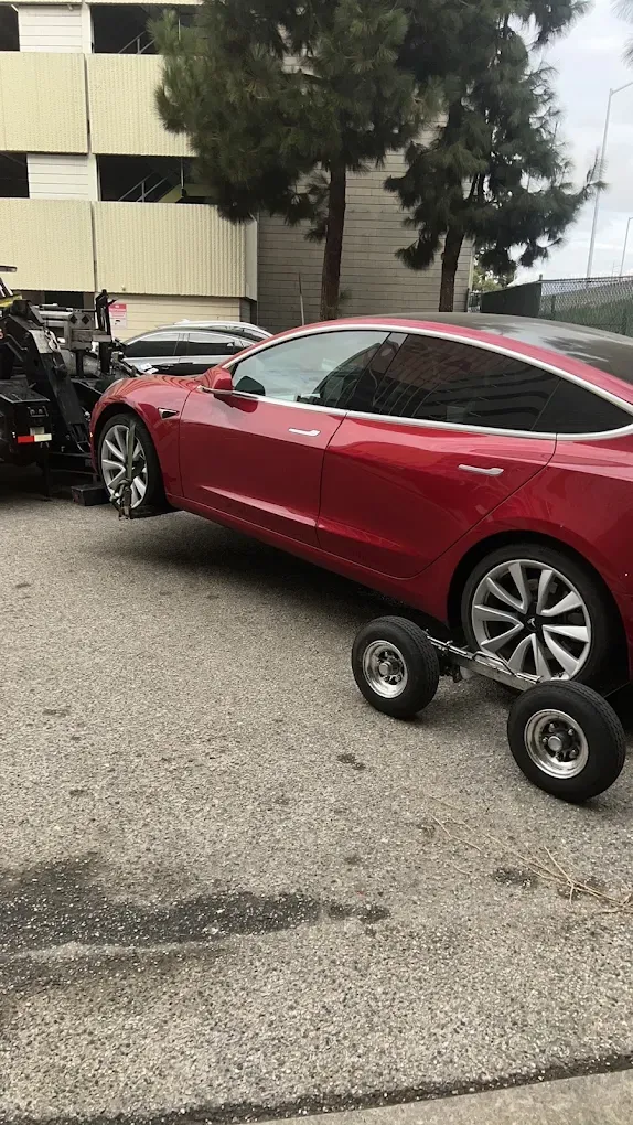 Red Tesla on wheel dollies behind an Urgent Towing truck
